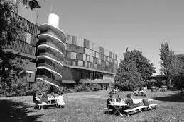 People sit at a bench on the grass by a statue and overlooking buildings